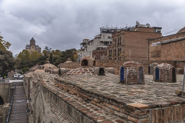A historic bathhouse with several domed brick structures is visible in the foreground, surrounded by a cityscape. In the background, there is a mix of older brick buildings and more modern constructions. A church with a pointed steeple is noticeable among trees, under a cloudy sky.