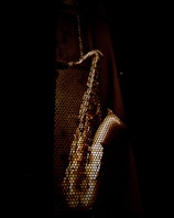 A saxophone is visible through a perforated metal surface, casting an artistic pattern of light and shadow. The background is dark, highlighting the metallic texture of the instrument.