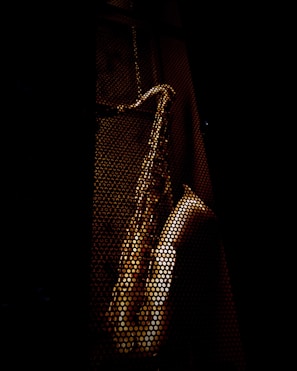 A saxophone is visible through a perforated metal surface, casting an artistic pattern of light and shadow. The background is dark, highlighting the metallic texture of the instrument.