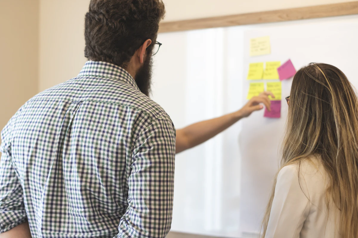 A woman placing pink and yellow sticky notes on a bright wall while a colleague stands beside her