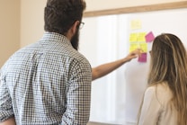 Two people are standing in front of a whiteboard covered with colorful sticky notes. One person is pointing at the notes, suggesting a collaborative work or brainstorming session.