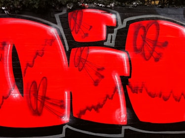 The image features a vibrant piece of graffiti art. Bold red and black colors create a striking visual impact. The design includes large, blocky letters with black outlines and smaller black doodles overlaid on the letters. The background is a dark wall, contributing to the vivid contrast.