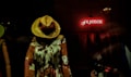 A mannequin dressed in a floral-patterned bohemian dress and wearing a wide-brimmed straw hat is displayed in a dimly lit window. In the background, a red neon sign spells out the word 'Freedom'. The scene gives off a vintage vibe with an emphasis on artistic and free-spirited expression.
