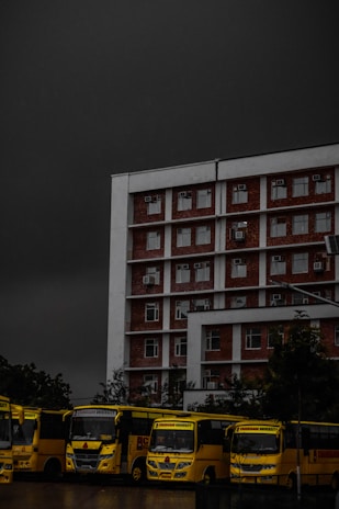A multi-story brick and white building is positioned prominently under a dark, ominous sky. In front of the building, a row of yellow buses is parked, lined up closely together. The buses have the text 'Chandigarh University' visible on them. Trees are partially visible in the background, adding some greenery to the scene.