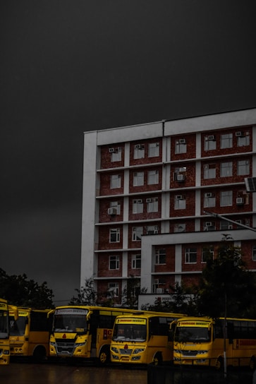 A multi-story brick and white building is positioned prominently under a dark, ominous sky. In front of the building, a row of yellow buses is parked, lined up closely together. The buses have the text 'Chandigarh University' visible on them. Trees are partially visible in the background, adding some greenery to the scene.