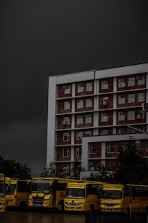 A multi-story brick and white building is positioned prominently under a dark, ominous sky. In front of the building, a row of yellow buses is parked, lined up closely together. The buses have the text 'Chandigarh University' visible on them. Trees are partially visible in the background, adding some greenery to the scene.