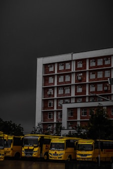 A multi-story brick and white building is positioned prominently under a dark, ominous sky. In front of the building, a row of yellow buses is parked, lined up closely together. The buses have the text 'Chandigarh University' visible on them. Trees are partially visible in the background, adding some greenery to the scene.
