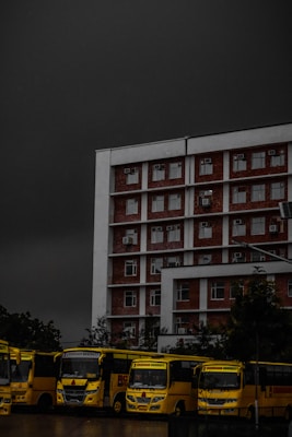 A multi-story brick and white building is positioned prominently under a dark, ominous sky. In front of the building, a row of yellow buses is parked, lined up closely together. The buses have the text 'Chandigarh University' visible on them. Trees are partially visible in the background, adding some greenery to the scene.