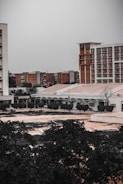 An urban landscape with multiple buildings, some tall and modern with a brick facade, and others shorter with a more utilitarian design. Trees and vegetation occupy the foreground, adding a touch of greenery to the otherwise muted architectural scene.