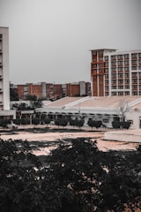 An urban landscape with multiple buildings, some tall and modern with a brick facade, and others shorter with a more utilitarian design. Trees and vegetation occupy the foreground, adding a touch of greenery to the otherwise muted architectural scene.