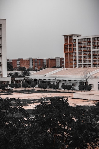 An urban landscape with multiple buildings, some tall and modern with a brick facade, and others shorter with a more utilitarian design. Trees and vegetation occupy the foreground, adding a touch of greenery to the otherwise muted architectural scene.