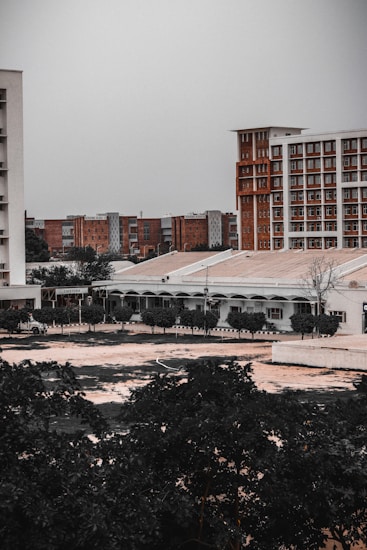 An urban landscape with multiple buildings, some tall and modern with a brick facade, and others shorter with a more utilitarian design. Trees and vegetation occupy the foreground, adding a touch of greenery to the otherwise muted architectural scene.