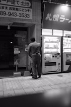 A person wearing a uniform stands in front of a vending machine on a city street. The scene is captured in black and white, adding a subdued and nostalgic atmosphere. A number of signs with Japanese text are visible, suggesting a location in Japan. The tiles on the ground and the urban setting indicate it's likely nighttime.