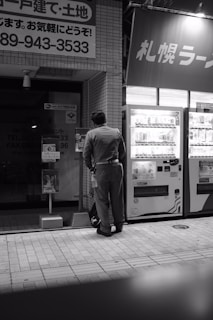 A person wearing a uniform stands in front of a vending machine on a city street. The scene is captured in black and white, adding a subdued and nostalgic atmosphere. A number of signs with Japanese text are visible, suggesting a location in Japan. The tiles on the ground and the urban setting indicate it's likely nighttime.
