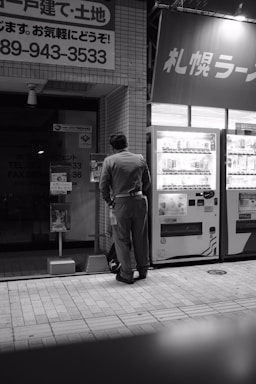 A person wearing a uniform stands in front of a vending machine on a city street. The scene is captured in black and white, adding a subdued and nostalgic atmosphere. A number of signs with Japanese text are visible, suggesting a location in Japan. The tiles on the ground and the urban setting indicate it's likely nighttime.