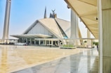 A large, modern mosque with a distinctive geometric design and a blend of traditional and contemporary architectural elements. Tall minarets flank the mosque, and there is a spacious courtyard with people walking around.