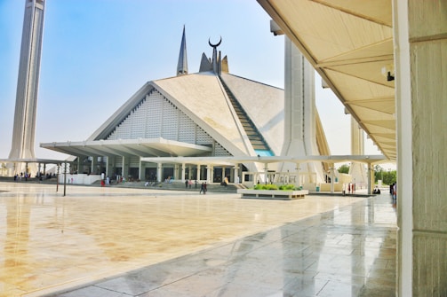 A large, modern mosque with a distinctive geometric design and a blend of traditional and contemporary architectural elements. Tall minarets flank the mosque, and there is a spacious courtyard with people walking around.