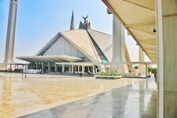 A large, modern mosque with a distinctive geometric design and a blend of traditional and contemporary architectural elements. Tall minarets flank the mosque, and there is a spacious courtyard with people walking around.