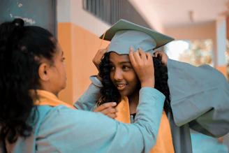 Two people are present, one assisting the other in adjusting a graduation cap and gown. Both are wearing academic robes with yellow stoles. They appear to be in an educational setting, possibly a hallway, with blurred background details.