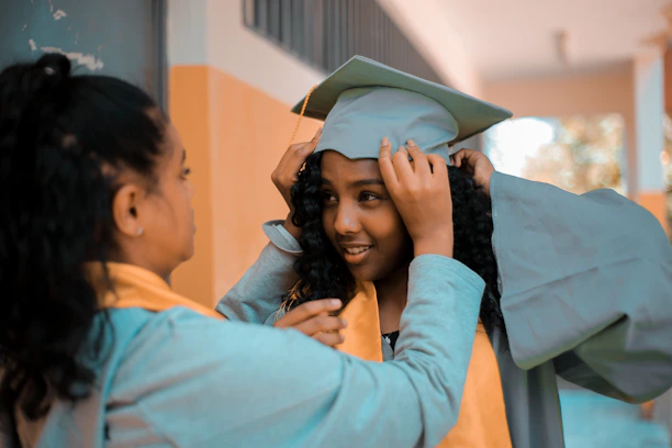 Two people are present, one assisting the other in adjusting a graduation cap and gown. Both are wearing academic robes with yellow stoles. They appear to be in an educational setting, possibly a hallway, with blurred background details.