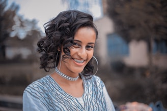 A young person with curly hair and braces is smiling warmly. They are wearing large hoop earrings and a patterned top with waves, complemented by a choker necklace. The background is blurred, consisting of trees and an indistinct building.
