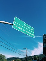 A green road sign is suspended in the air against a clear blue sky. The sign contains directions to Centro and Lagoa da Conceição, with arrows pointing in different directions. Below the sign, telephone poles and electrical wires stretch across the scene, with a hint of greenery and rooftops visible in the background.