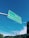A green road sign is suspended in the air against a clear blue sky. The sign contains directions to Centro and Lagoa da Conceição, with arrows pointing in different directions. Below the sign, telephone poles and electrical wires stretch across the scene, with a hint of greenery and rooftops visible in the background.