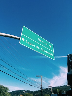 A green road sign is suspended in the air against a clear blue sky. The sign contains directions to Centro and Lagoa da Conceição, with arrows pointing in different directions. Below the sign, telephone poles and electrical wires stretch across the scene, with a hint of greenery and rooftops visible in the background.