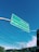 A green road sign is suspended in the air against a clear blue sky. The sign contains directions to Centro and Lagoa da Conceição, with arrows pointing in different directions. Below the sign, telephone poles and electrical wires stretch across the scene, with a hint of greenery and rooftops visible in the background.