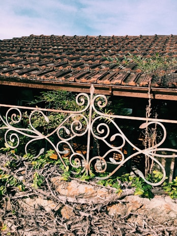 A rustic roof made of clay tiles sits above a decorative metal railing with intricate scrollwork patterns. Below the railing, there is a stone wall adorned with climbing green plants and vines. The scene is bright and sunny, with a clear blue sky in the background.