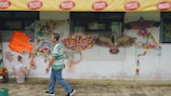 A man wearing a green striped shirt and blue jeans walks past various colorful kites displayed against a wall. The kites are of different shapes, including a fish, butterfly, and bird. Vibrant pinwheels and decorations are also visible near the kites. A yellow awning with red text advertising Vitasoy is above.