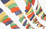 Brightly colored flags are strung across a light background, with a combination of red, green, yellow, blue, and orange hues. The flags are arranged in tightly packed, staggered rows, creating an overlapping pattern as they sway gently.