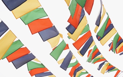 Brightly colored flags are strung across a light background, with a combination of red, green, yellow, blue, and orange hues. The flags are arranged in tightly packed, staggered rows, creating an overlapping pattern as they sway gently.