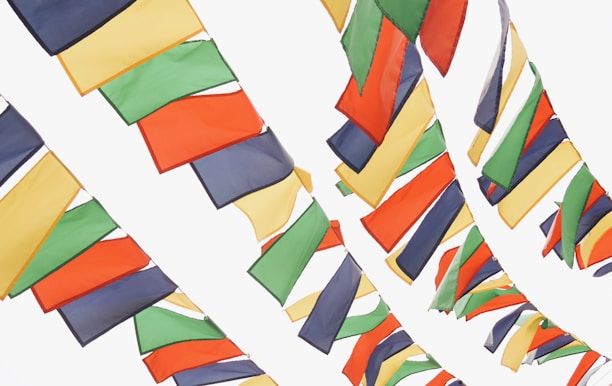 Brightly colored flags are strung across a light background, with a combination of red, green, yellow, blue, and orange hues. The flags are arranged in tightly packed, staggered rows, creating an overlapping pattern as they sway gently.