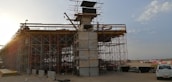 An under-construction bridge with scaffolding and concrete pillars, situated in an open area with clear skies. A crane and various construction materials are visible, with a vehicle parked nearby.