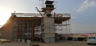 An under-construction bridge with scaffolding and concrete pillars, situated in an open area with clear skies. A crane and various construction materials are visible, with a vehicle parked nearby.