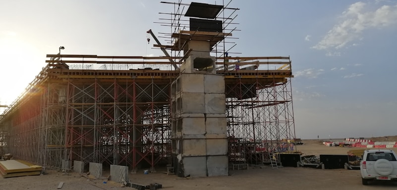 An under-construction bridge with scaffolding and concrete pillars, situated in an open area with clear skies. A crane and various construction materials are visible, with a vehicle parked nearby.