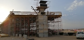 An under-construction bridge with scaffolding and concrete pillars, situated in an open area with clear skies. A crane and various construction materials are visible, with a vehicle parked nearby.