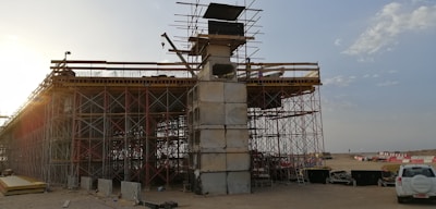 An under-construction bridge with scaffolding and concrete pillars, situated in an open area with clear skies. A crane and various construction materials are visible, with a vehicle parked nearby.