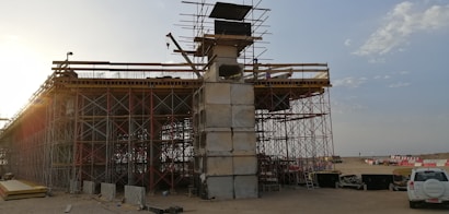 An under-construction bridge with scaffolding and concrete pillars, situated in an open area with clear skies. A crane and various construction materials are visible, with a vehicle parked nearby.