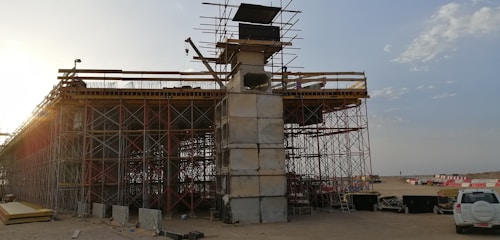 An under-construction bridge with scaffolding and concrete pillars, situated in an open area with clear skies. A crane and various construction materials are visible, with a vehicle parked nearby.