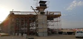 An under-construction bridge with scaffolding and concrete pillars, situated in an open area with clear skies. A crane and various construction materials are visible, with a vehicle parked nearby.