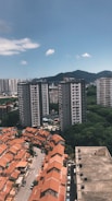 The image depicts a cityscape with a mix of residential buildings. In the foreground are rows of houses with orange roofs, and in the background are several tall apartment buildings. There is a hill or mountain covered with greenery in the background, under a clear blue sky with a few scattered clouds.