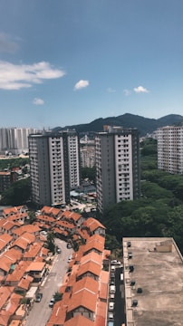 The image depicts a cityscape with a mix of residential buildings. In the foreground are rows of houses with orange roofs, and in the background are several tall apartment buildings. There is a hill or mountain covered with greenery in the background, under a clear blue sky with a few scattered clouds.