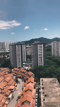 The image depicts a cityscape with a mix of residential buildings. In the foreground are rows of houses with orange roofs, and in the background are several tall apartment buildings. There is a hill or mountain covered with greenery in the background, under a clear blue sky with a few scattered clouds.