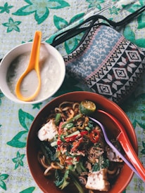 A vibrant dish of noodles with various toppings, including sliced red chilies, green vegetables, and tofu, served in a brown bowl. Beside it is a bowl containing a white creamy soup with an orange spoon. The tablecloth is decorated with green floral patterns, and a woven pouch with geometric designs is placed nearby.