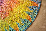 A vibrant mosaic artwork rich with geometric patterns in an array of colors including yellow, orange, red, and blue. The tiles vary in shape and size, creating a dynamic and textured appearance. Circular tiles are scattered throughout the design, adding depth and contrast.