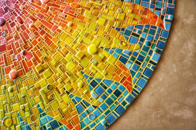 A vibrant mosaic artwork rich with geometric patterns in an array of colors including yellow, orange, red, and blue. The tiles vary in shape and size, creating a dynamic and textured appearance. Circular tiles are scattered throughout the design, adding depth and contrast.