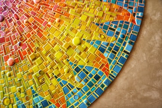 A vibrant mosaic artwork rich with geometric patterns in an array of colors including yellow, orange, red, and blue. The tiles vary in shape and size, creating a dynamic and textured appearance. Circular tiles are scattered throughout the design, adding depth and contrast.