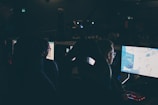 Several individuals are sitting in front of computer screens in a dimly lit room, likely engaging in gaming or working on tasks. The glow of the monitors illuminates their faces, creating a focused and immersive atmosphere. The surroundings suggest a setting like a gaming event or a LAN party.